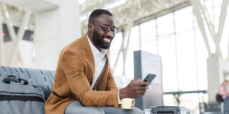 Man in a brown jacket sits on an airport bench, smiling at his phone while updating his T7E expenses. A coffee cup and suitcase sit beside him as the bustling, modern terminal unfolds in the bright background.