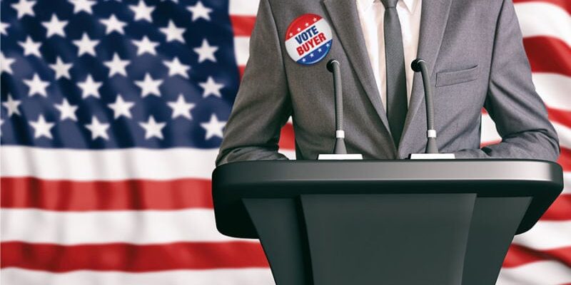 A person in a suit stands at a podium with microphones, wearing a "Vote Buyer" button. The background features the American flag.
