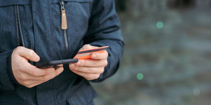 Person in a dark jacket holds a smartphone and a credit card, poised for online payment, while embracing the ease of automated reconciliation.