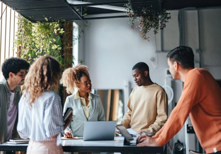 A group of five people stands around a table with a laptop, engaged in discussion about streamlining back office operations through Ai and automation inside a room with hanging plants.
