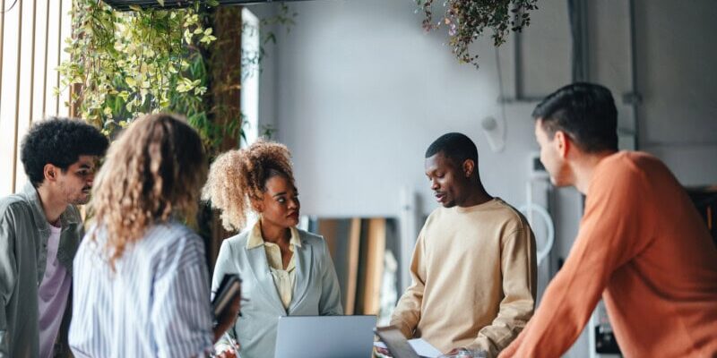 A group of five people stands around a table with a laptop, engaged in discussion about streamlining back office operations through Ai and automation inside a room with hanging plants.