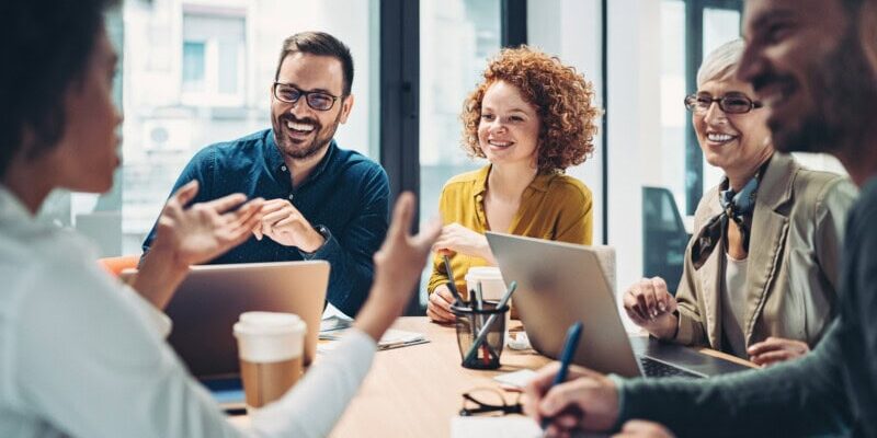 Five people sit at a table in a modern office, engaged in discussion with laptops, notebooks, and coffee cups in front of them.