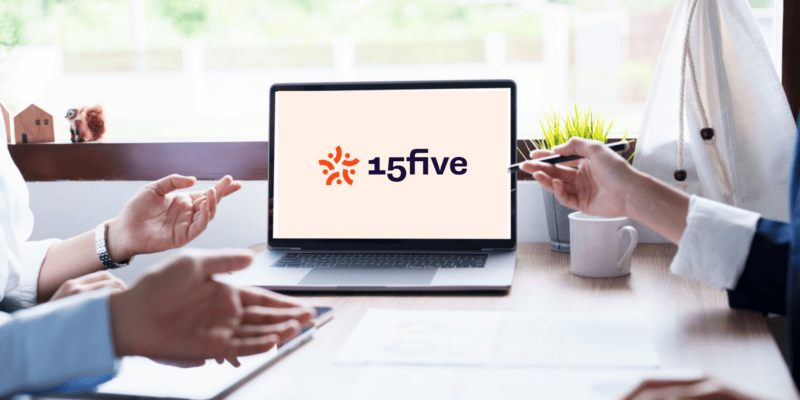 Two people sit at a table gesturing in discussion. A laptop displaying the "15five" logo is on the table. A pen is held by one person, a plant and coffee cup are nearby.