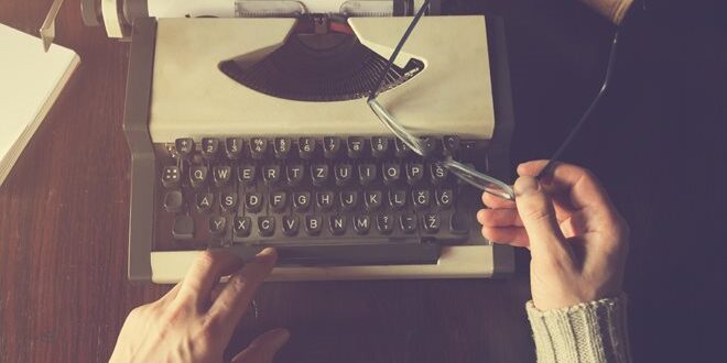Person typing on a vintage typewriter, holding glasses in the other hand, with a book nearby.