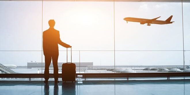 A person in a suit stands with a suitcase, gazing at a plane taking off through a large airport window.