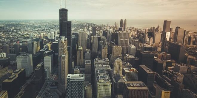 Aerial view of a sprawling cityscape with numerous skyscrapers under a cloudy sky, featuring a prominent tall building in the foreground.