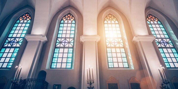 Interior of a church with arched stained glass windows, sun streaming in. Candles and framed artworks are mounted on the walls.