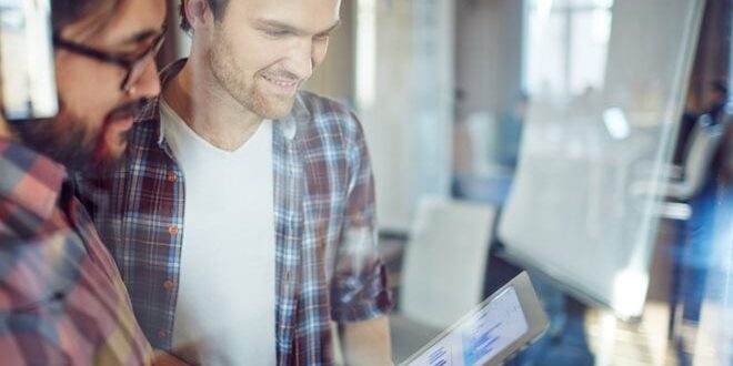 Two men in a bright office looking at a tablet displaying charts, with a flip chart in the background.
