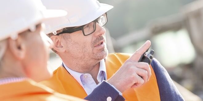 Two construction workers in helmets and orange vests are talking. One is pointing, and the other is partially visible.