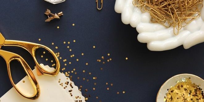A flat lay of gold scissors, paper clips, binder clips, and scattered confetti on a dark surface, alongside a bowl with more confetti.