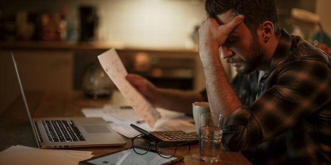 A man sits at a table with a laptop and paperwork, holding his head in frustration.