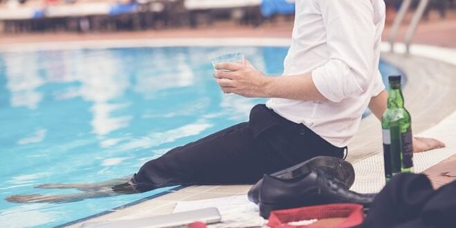 Man in a white shirt and black pants sits by a pool, holding a drink. Nearby are a bottle, laptop, and red bag.
