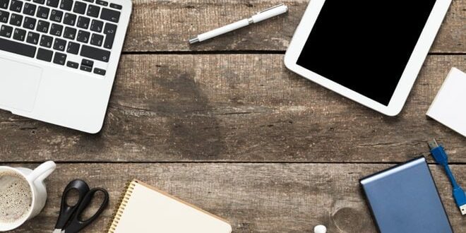 Wooden desk with a laptop, tablet, smartphone, notebook, pen, glasses, earbuds, scissors, coffee mug, external drive, and charging cables.