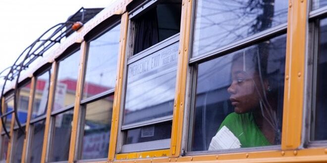 Person in a green shirt looks out the window of a yellow school bus.