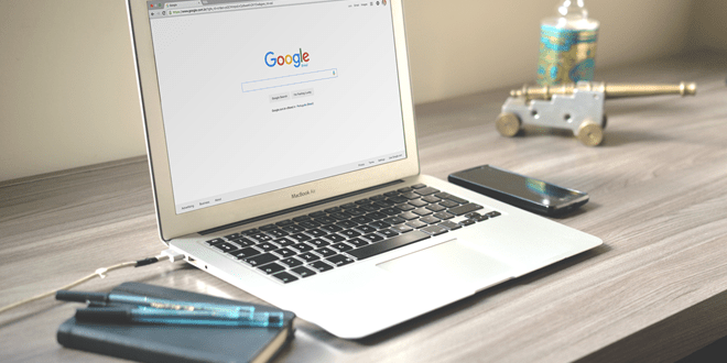 A laptop on a desk displaying the Google homepage, with a smartphone, notebook, pen, and decorative bottle nearby.