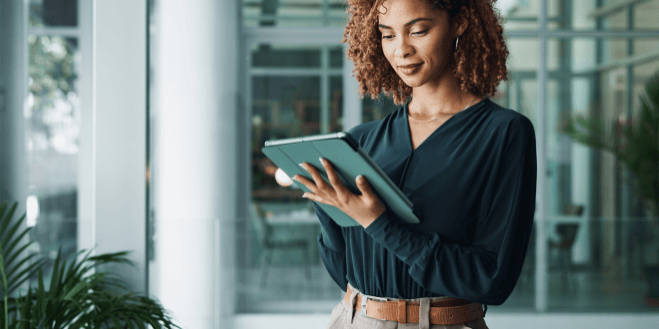 With curly hair framing their face, a person delves into a tablet, embodying the efficiency of back office operations against a backdrop of greenery and glass doors.