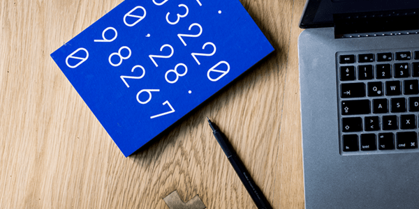 Blue notebook with white numbers, black pen, and a laptop on a wooden desk.