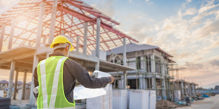 A construction worker in a yellow hard hat and safety vest reviews blueprints at a building site with partially completed structures and scaffolding under a blue sky.