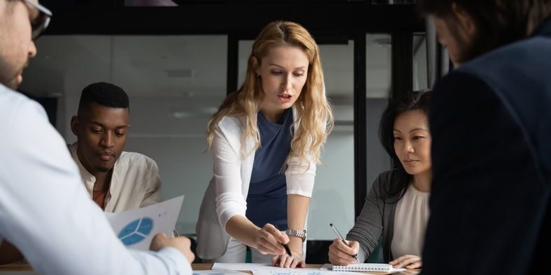A group of five people are in a meeting room discussing papers with charts on a table. A woman in the center is pointing at a document.