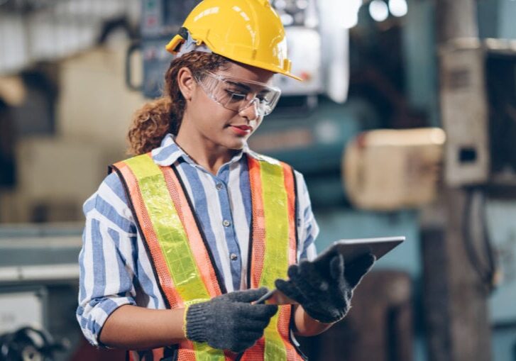 A woman in safety gear, including a yellow helmet and safety vest, uses a tablet in an industrial setting.