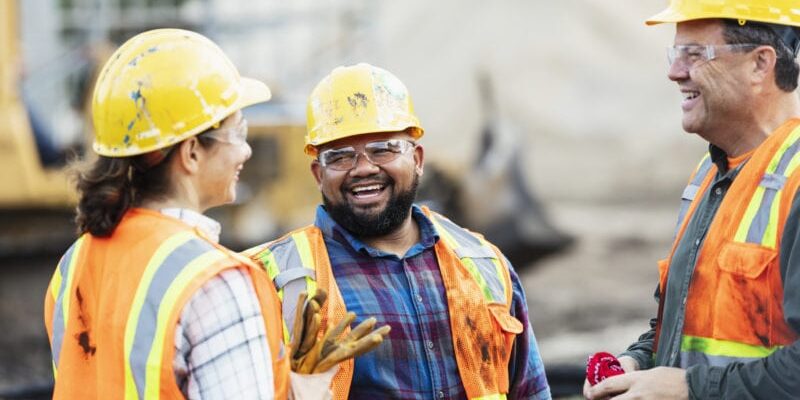 Three construction workers in hard hats and safety vests are smiling and talking on a construction site.