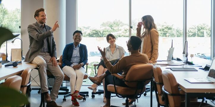 A diverse group of five people are having a lively discussion in a bright office space, seated around a table with laptops and papers.