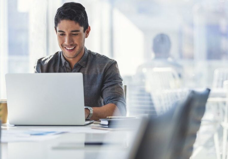 A person sits in a bright office, smiling and working on a laptop.