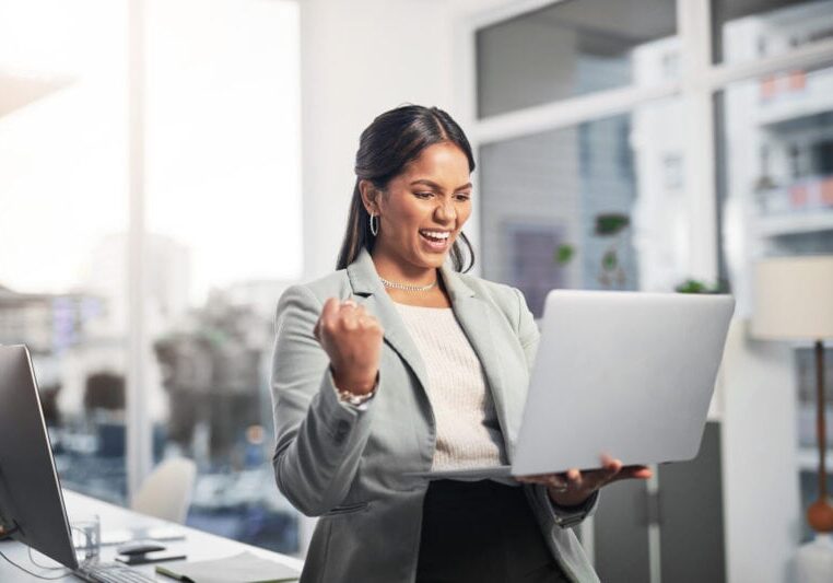 A person in business attire smiles and pumps their fist while looking at a laptop in a bright office.