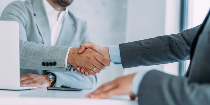Two people in business attire shaking hands across a table, with a laptop and a smartphone nearby.