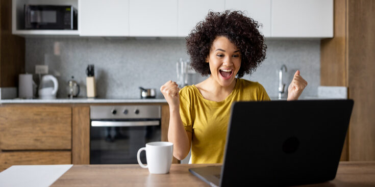 A person with curly hair looks excited while sitting at a kitchen table with a laptop and a coffee mug.