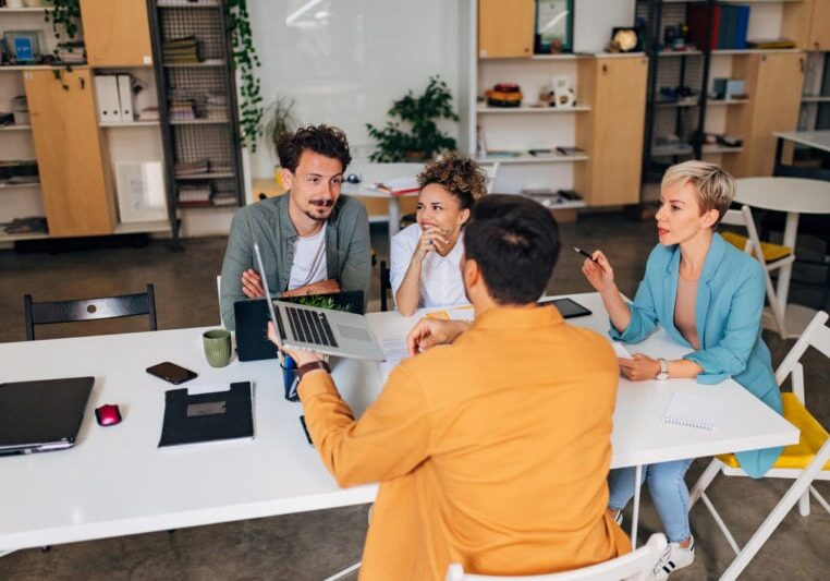 Four people sit around a table in a modern office, discussing and viewing a laptop.