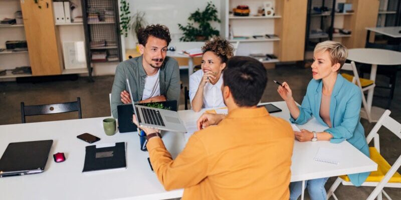 Four people sit around a table in a modern office, discussing and viewing a laptop.