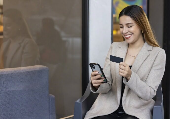 A woman in a beige blazer sits and smiles at her phone while holding a prepaid card.