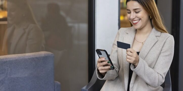 A woman in a beige blazer sits and smiles at her phone while holding a prepaid card.