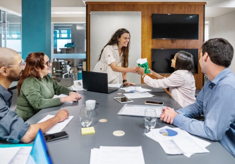Five people in a meeting room, seated around a table with documents and laptops, while one person hands a gift bag to another.