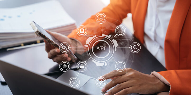 Person in an orange blazer uses a smartphone and laptop. A virtual diagram labeled "API" features icons like a lock, gear, and cloud, symbolizing technology and connectivity concepts.