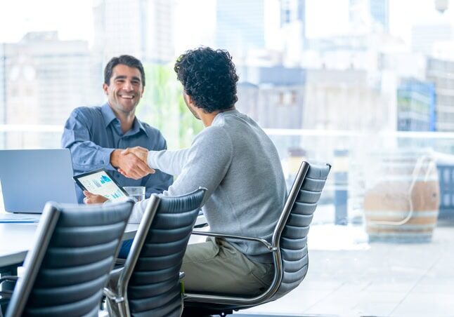 Two men shake hands across a conference table in a modern office with large windows and city views. One is holding a tablet displaying charts.