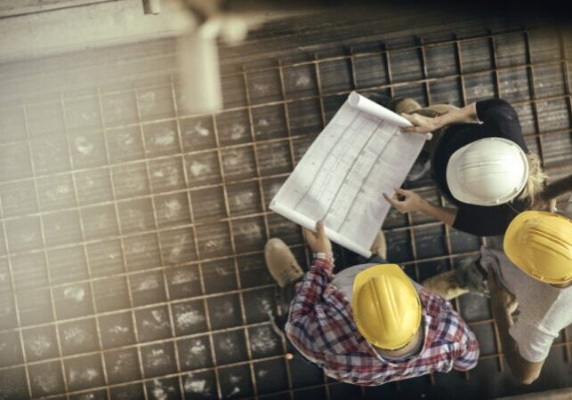 High angle view of three people with helmets, female architect, foreman and engineer on a construction site, looking down on a blueprint, copy space.