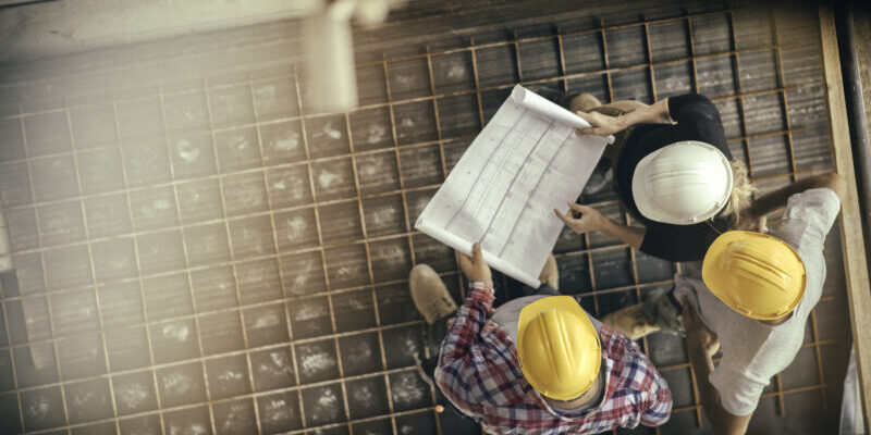 High angle view of three people with helmets, female architect, foreman and engineer on a construction site, looking down on a blueprint, copy space.