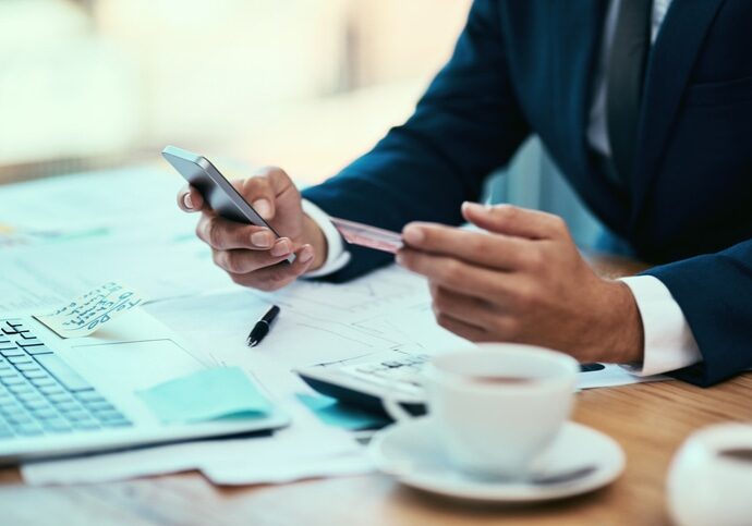 A person in a suit holding a smartphone and credit card at a desk with a laptop, documents, and a coffee cup.