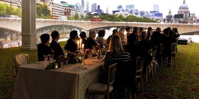 A group of people sit at a long dining table in a room with large projector screens displaying a cityscape with a bridge and skyline, creating an immersive dining experience.