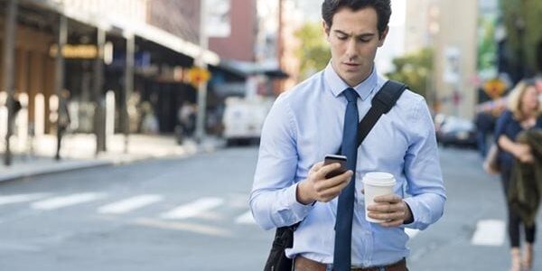 A man in a blue shirt and tie walks on a city street, holding a smartphone and a coffee cup. He has a bag slung across his shoulder.