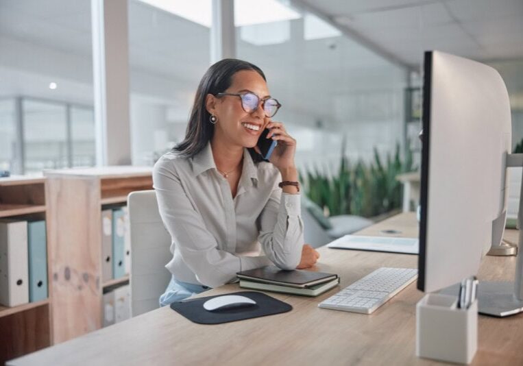 A woman sits at a desk in a modern office, smiling while talking on a mobile phone, with a computer monitor, keyboard, and notebooks in front of her.