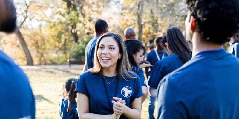 A group of people wearing matching blue shirts are gathered outdoors, talking about their nonprofit expenses and smiling, with trees and sunlight in the background.