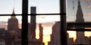 Blurred city skyline at sunset viewed through window with partially closed blinds.
