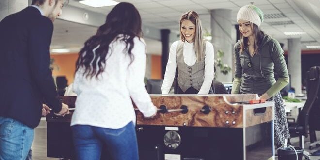 Four people playing table football in an office setting, with modern decor and open workspace in the background.