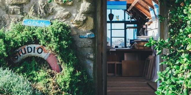 Entrance to a stone-walled studio surrounded by greenery, with a view of the interior featuring wooden shelves and a blue open sign above the doorway.