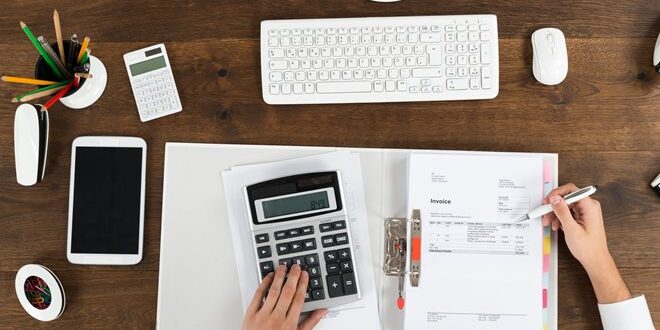 Person working at a desk with a calculator, keyboard, tablet, and invoices.