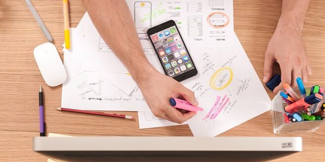 Person marking design sketches with a pink highlighter, surrounded by a smartphone, computer, mouse, and colorful pens on a wooden desk.