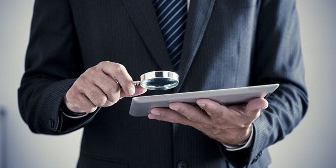 A person in a suit examines a tablet closely with a magnifying glass.